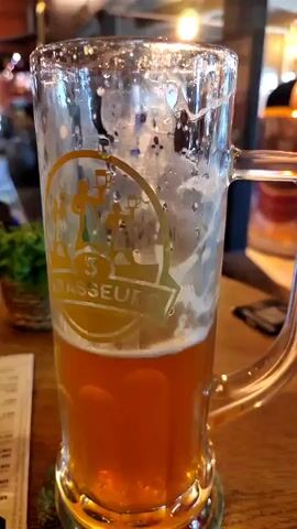 The video shows a close-up of a glass mug filled with amber-colored beer. The mug has a logo on it that appears to be of three figures holding steins. The background is blurred, but it seems to be a brewery or a bar with industrial-style decor. There are copper brewing vats visible, as well as exposed brick and wooden elements. The lighting is warm and inviting. The camera pans slightly, giving a wider view of the establishment. There are tables and chairs in the background, suggesting a seating area. In one shot, a spiral staircase is visible. The overall impression is a modern, yet rustic, brewery environment.