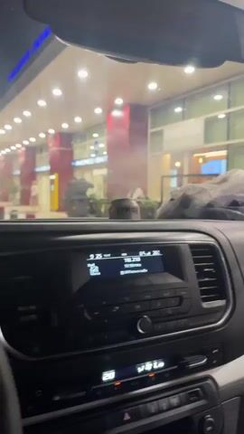 The video begins inside a car, showing the dashboard and the view through the windshield of a brightly lit building with red architectural elements and green plants. A man in a white cap, denim jacket, and blue fanny pack opens the front passenger door and stands outside. Several people gather at the car's open door, including a woman in a yellow dress with glasses and a woman in a white flowing top with a studded bag. The camera then shifts inside the car, showing the woman in the white top and another woman with dark hair in a dark shirt entering the back seat. The woman in the white top leans forward in the back seat, while the woman in the dark shirt is also visible.