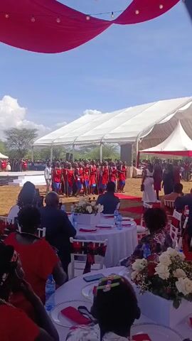 The video depicts an outdoor wedding celebration. Under a large white tent adorned with red drapery, seated guests observe a group of people in traditional red and blue clothing holding sticks, presumably performing a cultural dance. The guests sit at tables decorated with white linens, floral centerpieces, and water bottles. A red carpet leads to the performance area. Beyond the tent, a landscape of trees and a partly cloudy sky are visible.