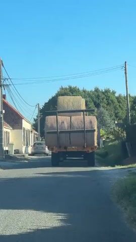 The video shows a rear view of a truck carrying large round bales of hay. The truck is driving down a paved road with trees lining both sides. On the left side of the road, there is a white house with a car parked in front of it. The sky is clear and blue. The truck has a yellow trailer with a metal railing around the hay bales. The hay bales appear to be stacked two high.