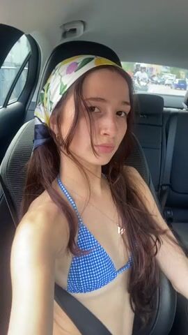 A young woman with long, dark brown hair and a blue and white gingham bikini top is seated in the passenger seat of a car. She is wearing a colorful silk scarf around her head, tied at the back with navy blue ribbons. The scarf has a pattern of yellow lemons and white and purple flowers. A delicate silver necklace with a letter "W" pendant is visible on her chest. Her lips are pursed, and she is looking directly at the camera with a playful expression.

The scene is set inside a car, with the dark interior of the vehicle visible. Through the windows, a blurry street scene can be seen, suggesting the car is either parked or moving slowly. The background shows other vehicles and buildings, indicating an urban or semi-urban environment. The lighting suggests it is daytime, likely a sunny day, as indicated by the bright light filtering into the car. The woman's relaxed posture and attire (bikini and scarf) suggest a casual outing, possibly related to a beach or a warm-weather destination. The overall mood is lighthearted and relaxed.