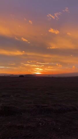 The video shows a wide, dark field at sunset. In the distance, a small building stands silhouetted. Beyond the field are rolling hills and a distant cityscape. The sky is filled with clouds colored in hues of orange, yellow, and purple from the setting sun.