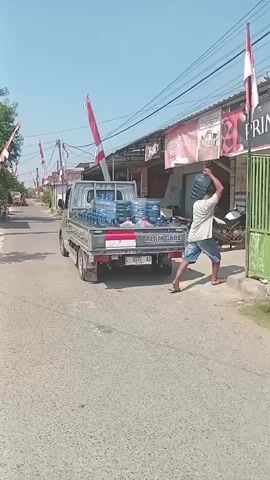A man is seen carrying a large water jug on his shoulder while walking towards a building. In front of the building, a light-colored pickup truck is parked, loaded with many similar water jugs. The truck has Indonesian flags attached to its sides. The street is paved, and there are shops and houses visible in the background. The sky is clear and blue. Another person is visible in the driver's seat of the truck. A motorcycle is parked near the building.