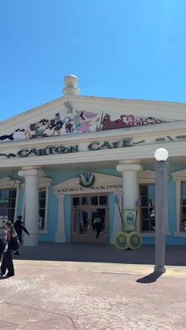The video shows a bright and sunny day at a theme park, likely one dedicated to Warner Bros. cartoons. Several people are seen walking around the park, including a group of teenagers walking in front of a building labeled "CARTOON CAFE" with classic cartoon characters displayed on the facade. The video also shows a large fountain with cartoon characters playing instruments and an archway entrance to an area labeled "LOONEY TUNES PARK." The overall atmosphere is lively and family-friendly, with colorful buildings and themed decorations.