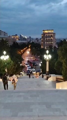 The video opens with a view looking down a wide, stone staircase. Two people are walking up the stairs from the bottom. On the left, a person wearing a dark jacket and dark pants is walking. On the right, a person wearing a white hooded jacket and lighter pants is walking. Both are facing up the stairs. At the bottom of the stairs, there is a street with several cars and buses driving. There are streetlights illuminating the road and sidewalks. Trees line the street and the area around the staircase. In the background, a cityscape is visible with several buildings, some of which are lit up. The sky is dark and cloudy, suggesting it is dusk or evening. As the video progresses, the camera pans to the right, revealing more of the cityscape. Three flame-shaped skyscrapers become prominent in the background, illuminated by artificial lights. Some of the buildings and trees in the middle ground are also lit with colored lights, including purple. The staircase continues to be visible in the foreground, with decorative lighting along its sides. The video focuses on the architectural elements of the city and the approach to an elevated vantage point.