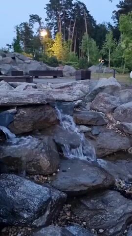 The video shows a man-made waterfall flowing over rocks. In the background, there are several large rocks, benches, trees, and lights, suggesting an outdoor park or garden setting. The sky is visible and appears to be twilight or early evening.