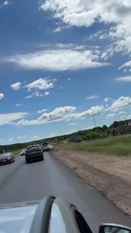 The video shows a view from a car driving on a road with other cars in front. The sky is blue with white clouds. The road is paved and appears wet in some areas. There are trees and green fields on the sides of the road. In the distance, there are some signs and a small building. The video is shot from a low angle, showing the roof of the car in the foreground. The camera is moving forward, indicating that the car is in motion.