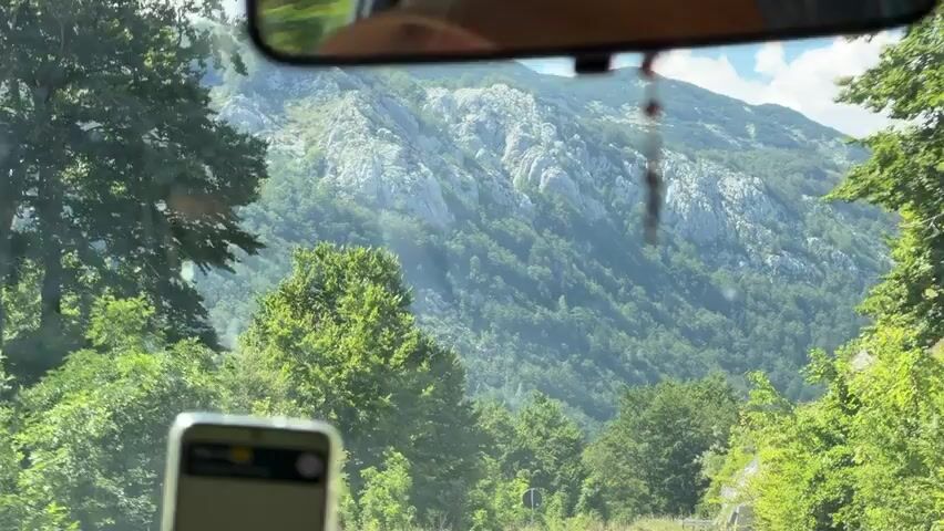 The video shows a view from inside a car looking out through the windshield. The car is driving on a road that curves to the right. On the right side of the road, there is a tall concrete wall. Beyond the wall, there are green trees and a steep, rocky mountain range in the background. The sky is blue with some white clouds. In the foreground, a smartphone is visible on the dashboard, displaying what appears to be a navigation app. Further down the road, a small orange car is visible. A road sign indicating no overtaking is also visible on the right side of the road.
