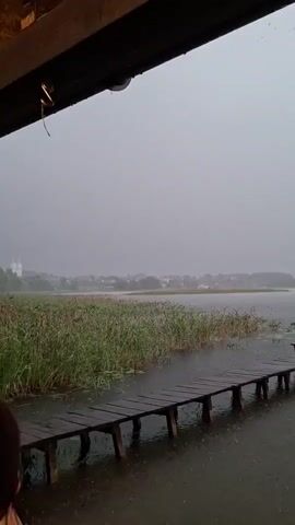 The video captures a scene of a lake during a rainstorm, viewed from under a wooden overhang. A wooden pier extends into the water on the right side of the frame. Tall reeds and grass line the edge of the lake. In the distance, a town or village with white buildings and steeples is visible across the water. The sky is overcast and gray, and the rain is visible falling onto the pier and the water. The sound of rain and distant thunder can be heard.