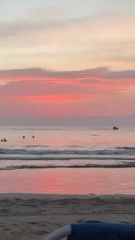 The video shows a beach scene at sunset or sunrise. The sky is filled with pink and orange hues, reflected in the calm water. Gentle waves are rolling onto the sandy beach. In the distance, there are a few people in the water, appearing as dark silhouettes. A jet ski briefly appears further out in the water. A blue object, possibly a beach chair, is partially visible in the foreground.