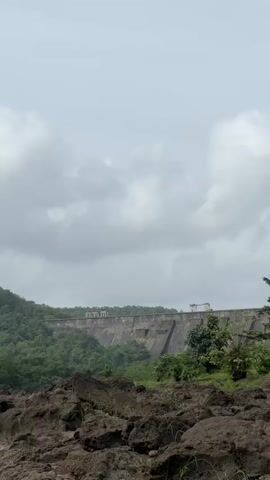 The video pans to the right, showing a large concrete dam stretching across the frame. The dam is surrounded by lush green hills and trees. The sky is overcast with scattered clouds. In the foreground, there are large piles of dark brown rocks and some green grass. The video captures a wide, scenic view of the dam and its natural surroundings.