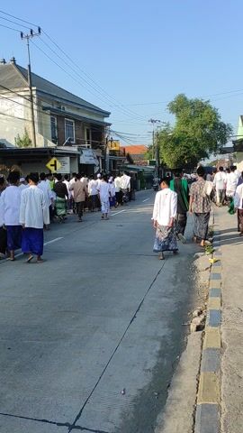 A procession of men and boys, primarily dressed in white shirts or robes and sarongs or pants, walks in an orderly line along a paved street. Many participants wear kufis or peci hats. Buildings, resembling houses and small businesses, line the left side of the road, featuring traditional Indonesian architecture. Utility poles and power lines are visible running parallel to the street. The sky is clear and blue, indicating bright, sunny weather. Long shadows cast across the road suggest either early morning or late afternoon. A yellow road sign with a black arrow pointing right is prominently displayed on the left side of the street. Another sign or banner is partially visible on one of the buildings further down the road. The individuals in the procession appear focused, participating in what seems to be a communal or religious event.