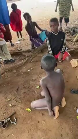 The video shows a group of children in an outdoor setting, possibly a village or rural area, with a dirt ground and tree roots visible. One boy is kneeling with his hands seemingly tied behind his back. Other children are standing or sitting around him, some looking at him. The children are wearing various clothing items. A metal object that looks like a lock or shackle is lying on the ground nearby. The overall atmosphere is unclear, but the kneeling boy's situation suggests a possible disciplinary action or a staged scene.