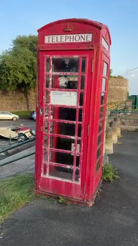 The video shows a traditional red British telephone booth. It is standing on a sidewalk with some greenery around its base. A hand with pink nail polish reaches out to touch the door.  The booth is weathered with peeling paint, and has signs inside, including a "No Smoking" sign and a notice that says "This kiosk is protected for future generations." Outside the booth there's a street with cars passing by, a stone wall, and a tree. The sky is clear and blue.