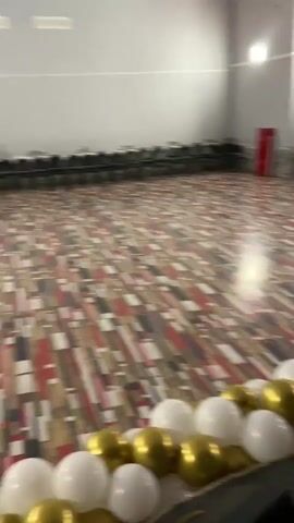 The video shows a decorated room with gold and white balloons. The room has multiple exits, and some seating is visible. The floors are a mix of carpet and wood. The lighting in the room appears to be standard overhead lighting. There are no people visible in the video.