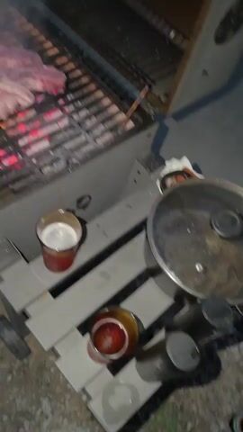 The video shows a person grilling meat over hot coals. The scene takes place outdoors at night, with the glow of the coals and flames illuminating the food and the grill. The meat appears to be steak or chops, and some pieces have been cut into strips. Smoke billows from the grill as the meat cooks. The person's hand is visible, holding a bottle and tilting it as if to pour liquid onto the grill, possibly to douse flames or add flavor. There are also some glasses visible on a shelf below the grill, one containing a light-colored beverage, suggesting a social gathering. The overall atmosphere appears relaxed and casual, typical of an outdoor barbecue. The location is specified as Slivo Pole, Bulgaria, though no specific landmarks are visible in the footage. The time of day is clearly night, indicated by the darkness of the surroundings and the focus on the illuminated grill. There are no visible signs or text in the video.
