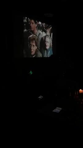 A video is projected onto a screen in a very dark room. The main subject of the projection is a man with dark hair and stubble, wearing a white shirt and red suspenders. He is holding a glass to his mouth and appears to be drinking from it. Faintly visible behind him on the screen are silhouettes of other people in what looks like a bar or performance setting.

Below the projected image, in the dark foreground of the room, several objects are dimly lit. There are some flat, rectangular items that resemble cards or papers scattered on a surface. To the right, a lit candle casts a warm glow. The overall setting suggests an intimate viewing experience, possibly a screening or performance, in a low-light environment. The audio includes music and speech, indicating that the projected video is part of a larger presentation. No specific text is visible in the video.