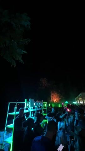 At night, an outdoor event features a bar with “Billboard” branding lit in green. A crowd gathers around it, their silhouettes visible against the bright bar and other light sources in the background.  Palm trees and other foliage punctuate the dark sky. In the background is a white structure, possibly a stage, with green lights highlighting its perimeter. The overall scene suggests a lively, possibly music-related, event.