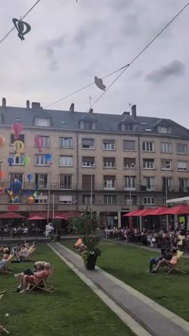 This is a vertical video of an outdoor public space. In the foreground, there are several people sitting in lounge chairs on a manicured lawn. A few more people are visible in the background, some sitting at tables outside of buildings. The buildings themselves are multi-story structures with many windows. A large number of colorful hot air balloon decorations are suspended from wires strung across the sky above the scene, some of which are catching the sunlight. The sky is overcast with patches of bright sun.