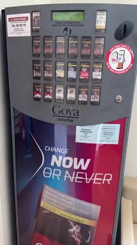 This media file captures a close-up view of a cigarette vending machine, likely located in Käerjeng, Luxembourg. The machine displays rows of cigarette packs, with brands such as Elixyr, Ducal, Camel, and Winston clearly visible. Some of the packs are in cellophane wrapping, indicating they are new and for sale.

The machine has a digital display at the top which shows messages in French and Spanish. At the beginning of the clip, it reads "PRIX 8.80 EUR INTRODUCE PIECE," indicating a price of 8.80 Euros and a prompt to insert payment. Later, the display changes to "VOTRE VISITE. INTRODUCE PIECE," suggesting a transaction has been initiated or completed, and a reminder to insert payment for a purchase. Below the display, there are instructions in Spanish and English: "PRECIO PULSANDO TECLA PRESS BUTTON TO DISPLAY PRICE."

A prominent graphic on the lower half of the machine features the slogan "CHANGE NOW OR NEVER" in large white lettering against a red background. Below this, there is health warning text in French and German, including "Fumer augmente le risque de devenir aveugle. Rauchen erhöht das Risiko zu erblinden" (Smoking increases the risk of becoming blind. Smoking increases the risk of blindness). A close-up of an Elixyr Red cigarette pack is also visible as part of this graphic.

A hand with manicured nails enters the frame from the right, reaching towards the cigarette selection area. This suggests a person is interacting with the machine, likely to make a purchase. The overall scene depicts a mundane commercial transaction at a vending machine, with a focus on the products being sold and the associated health warnings. The lighting appears to be indoor, consistent with a shop or public space.
