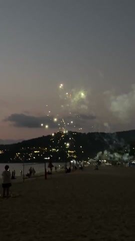The video depicts a beach at dusk, with a dimly lit coastline dotted with buildings visible in the background. Several people are visible, some standing near the water's edge, others sitting on the sand. There's smoke in the air, possibly from fireworks or other celebratory activities, and there's a series of sparks or lights visible in the sky.