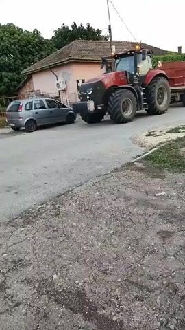 A red tractor is driving down a road. It is pulling a trailer. A man on a scooter is riding next to the tractor. The scooter is white and has a person on it wearing a white shirt and blue shorts. The road is paved and has gravel on the side. There are houses and trees in the background. The sky is cloudy.