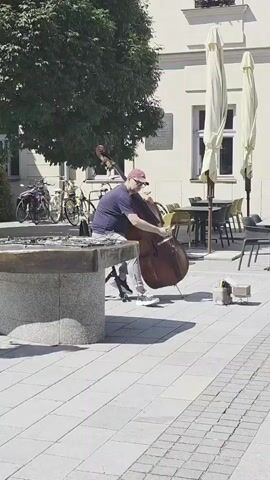 A lone man is playing a double bass in an outdoor square. He is wearing a blue t-shirt, grey shorts, a red baseball cap, and white sneakers. He is seated on a small black stool next to a stone structure, possibly a fountain, and is leaning over the instrument. To his right, on the paved ground, are a few small, brown boxes. In the background, there are tables and chairs from an outdoor cafe, two large white umbrellas, and a building with windows. To the left of the man, there are several parked bicycles. The scene is bathed in sunlight, casting shadows.