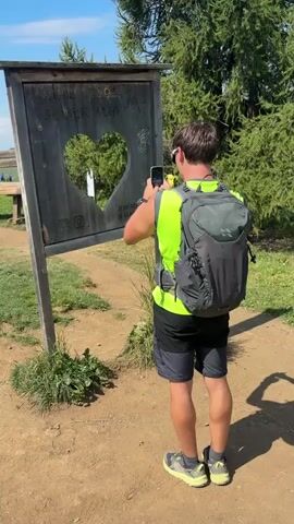 A young person wearing a neon yellow vest and a dark backpack stands with their back to the camera, holding a smartphone and taking a picture of a wooden frame with a heart-shaped cutout. The frame has writing on it that is partially obscured. Through the heart-shaped cutout, two other people can be seen in the distance, sitting on a bench. The environment appears to be outdoors, with trees and a clear blue sky visible. The ground is dirt. The person with the phone is looking at their phone screen and then appears to look through the heart cutout. The timecode indicates the video is less than 6 seconds long, showing a brief moment.