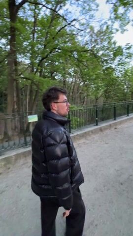 The video shows a man walking along a gravel path in what appears to be a park or green space. He's wearing a black puffer jacket, black pants, and glasses. The path is bordered by a low metal fence on one side, beyond which are trees. The sky is visible above the trees, appearing somewhat overcast. The camera follows the man as he walks, moving past him so that at one point, another individual is briefly visible.