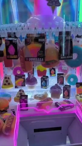 The video shows the inside of a claw machine filled with plush toys shaped like food items such as donuts, pizza slices, cupcakes, ice cream cones, burgers, and hot dogs. The toys are hanging from the top of the machine, and some are displayed on shelves below. The machine is brightly lit with colorful LED lights. At the top of the machine, a cartoon cat character wearing sunglasses is visible. The claw mechanism is at the top, ready to drop. The video appears to be taken from the front of the machine, looking in.