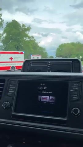 The video shows a road blocked by multiple vehicles, including at least two ambulances and a gray MAN van labeled “MEILENSTEIN TIEFBAU GMBH.” The ambulances are red and white with the German Red Cross emblem. An orange warning triangle is placed on the road. The van has its driver-side door open, and there appears to be debris scattered on the road surface near it. Traffic is backed up behind the incident. The video is shot from inside a vehicle, showing the dashboard and car’s infotainment system, which displays music information. The weather appears overcast, with green trees lining the sides of the road. The license plate on a gray Audi station wagon in front of the recording vehicle reads “VER-TS 33.” The sky is overcast.