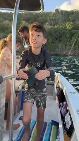 A young boy on a boat proudly holds up a freshly caught fish. He is wearing a black rash guard with "SURF" written in camouflage across the chest and camouflage shorts. The fish is dark blue with teal and yellow markings, and has a distinctive fin. The boy looks excited and happy.

In the background, a man with a beard is also on the boat, wearing a black long-sleeved shirt. Another person, possibly a girl, is visible behind the boy. The boat appears to be on the water, with lush green hills and trees forming the coastline in the distance. The sun is setting, casting a warm glow on the scene, suggesting it is late afternoon or early evening.

A fishing rod is visible extending from the back of the boat. The overall setting is a picturesque coastal area, likely a bay or inlet, with calm water. The mood is one of accomplishment and enjoyment of an outdoor activity. The visible text on the boy's shirt is "SURF".