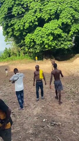 This video captures a vibrant outdoor scene in Unwana, Nigeria, featuring a group of young men and children on a reddish-brown dirt clearing. The setting is rural, characterized by large, lush green trees dominating the background and a bright blue sky with scattered clouds above. Power lines stretch across the frame, and a tall palm tree is visible in the distance.

The primary activity is dancing and celebratory movement. Several young men are actively engaged, displaying energetic and expressive dance moves. One young man in the foreground wears a yellow t-shirt with faces printed on it and text that appears to say "Unu Aseela." He is seen gesturing with his arms. Another man, shirtless with a yellow cloth draped over his shoulder, is smiling broadly as he moves. Other participants include a man in a light grey t-shirt and jeans, and another shirtless man in dark shorts, all exhibiting dynamic movements.

In the background, more children and adults are scattered around the clearing, some observing and others participating in the general atmosphere of celebration. The overall mood appears joyful, with visible smiles on several faces. The time of day is clearly daylight, indicated by the bright, natural lighting. The ground is unpaved, suggesting a community gathering space.
