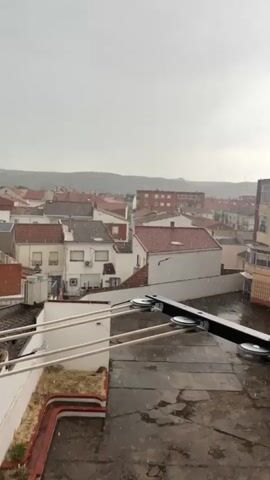 From a high vantage point, the scene captures a townscape under a light shower of rain. Predominantly, a collection of buildings with red tile roofs sprawls across the frame, punctuated by a few taller, apartment-style buildings with multiple windows. A prominent church steeple rises in the distance. In the foreground, a balcony with a clothesline system is visible, the metal components of which are damp from the rain. The overall lighting is subdued, reflecting the overcast sky. The environment is urban yet compact, suggesting a small or medium-sized town.