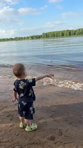 A young boy stands on a sandy beach, facing a body of water, possibly a river. He is dressed in a matching floral-patterned shirt and shorts, and wears light green Crocs. He is holding his arm out toward the water. A girl, who appears older, approaches him and places a hand on his shoulder. She is wearing a white crop top and black shorts, and is barefoot. The sky is blue with scattered clouds, and a line of trees can be seen in the background across the water.