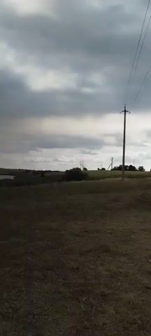 A video of a landscape with a cloudy sky. A power line runs diagonally across the top of the frame. In the foreground, dry grass covers the ground. In the middle ground, there is a pile of debris including branches and a green bag, and further back, a white SUV is parked. In the background, there are rolling hills covered in grass and trees, and a body of water on the left. There are no people visible in the video.