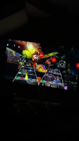 A person is playing Guitar Hero on a small TV screen. The background is dark. On the TV, a cartoon character stands on a stage and plays a guitar. The screen displays notes in various colors, which the player must match on the controller. Red Chinese lanterns hang in the virtual background.