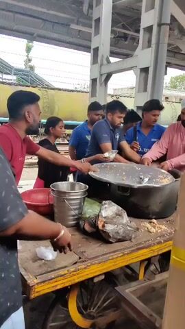 A group of men are serving food from a large pot on a cart at what appears to be a train station. The men are scooping food onto plates and handing them to people in line. The scene is bustling with activity, and there are several people standing around watching. The food appears to be rice-based, and it is being served with some type of condiment. There are also some large rocks or debris on the cart, as well as some wrapped items. A train is visible in the background.