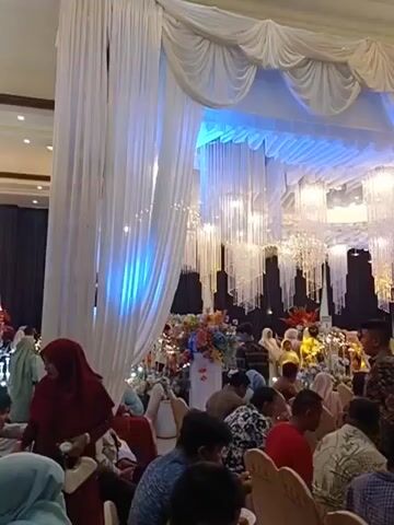 The video shows a wide shot of a large indoor event space, likely a banquet hall, decorated for a celebration. White drapes hang from the ceiling, some illuminated with blue light. Several large, ornate crystal chandeliers are suspended from the ceiling, along with numerous strands of smaller crystalline decorations. In the background, there is a stage area with more elaborate decorations, including floral arrangements and possibly a raised platform or altar. A crowd of people, mostly seated in chairs with beige covers and a logo that appears to read "NCH," are visible throughout the space. Many are dressed in formal or semi-formal attire, with some women wearing headscarves. The lighting is dim, with spotlights and ambient light creating a festive atmosphere. The focus is on the overall scene rather than any specific individual or action.