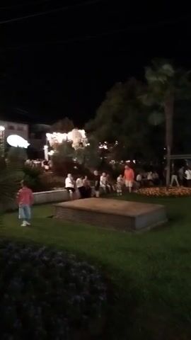 This is a shaky, nighttime video clip showing a park-like area with a grassy lawn and some trees. Several people are gathered in the background, some sitting and some standing. There are also buildings visible in the distance, with lights illuminating them and strung across the scene. In the foreground, a raised, rectangular object covered in metal is present. A person in a pink shirt and white shorts is in the lower left corner of the frame, and another person in a white dress or costume is standing in the middle ground. The video appears to capture an outdoor event or gathering at night.