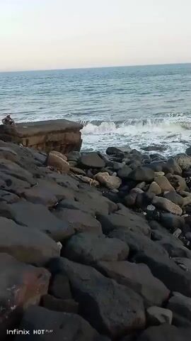 The video shows a rocky shoreline with waves crashing against the rocks. In the distance, there is a person sitting on a large rock. The sky is clear and the ocean is calm. The camera pans across the scene, showing the texture of the rocks and the movement of the waves. The video ends with a shot of the ocean and the sky.