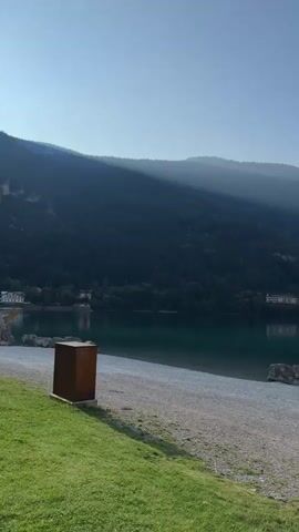 The video shows a still image of a scenic view of a lake with mountains in the background. The sun is shining, casting shadows on the mountainside. There is a grassy area in the foreground with a small, brown trash can. A gravelly beach runs along the edge of the lake, and a few rocks are visible in the water. The lake is a deep green color, and the sky is a clear blue with a few wispy clouds. The overall impression is one of peace and tranquility.