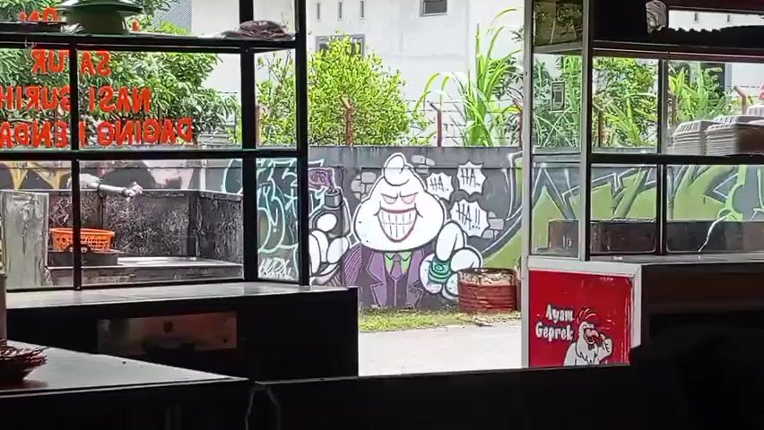 The video pans across a street-level view through a glass storefront. On the left, a metal cooking station with an orange basket is visible. In the center, a mural of a cartoon character resembling the Joker, with a white, round head, a large grin, and wearing a purple suit and green tie, is painted on a concrete wall. The character is holding a spray can in its left hand and money in its right. To the right, a red sign with a cartoon chicken and text that reads "Ayam Geprek" is displayed. The background features lush green foliage and buildings. The video appears to be shot from inside a shop looking outward.