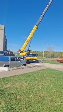 The video shows a construction area on a sunny day with a clear blue sky. A large yellow crane is positioned on the left side of the frame, its boom extending upwards. Several workers wearing orange vests are gathered near a stack of orange circular objects. In the background, a white flatbed truck is parked, and further to the right, a military tank is displayed on a platform. The ground is covered with green grass and paved walkways. The workers appear to be preparing for a lifting operation using the crane.