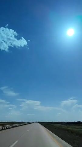 The video shows a POV shot from a vehicle traveling on a highway under a bright blue sky with a few scattered clouds. The sun is visible and appears very bright, casting a glare. The highway is empty except for a few cars in the distance. The landscape on either side of the road is flat and appears to be a mix of dry grass and some vegetation. The overall impression is a sunny day on a long road trip.