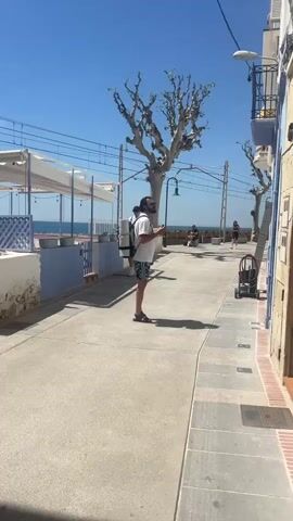 The video shows a man standing on a sunny promenade, eating a snack. He wears a white t-shirt, black and white patterned shorts, and dark sandals. He also has sunglasses and a backpack, with a rolled-up mat attached to it. The promenade has trees, benches, and buildings on either side. In the background, there's a view of the ocean. Other people can be seen walking and sitting on the promenade. The man stands in the middle of the frame, taking a bite of his snack and looking around.