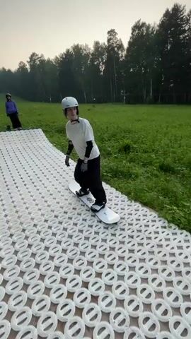 A person wearing a white t-shirt, black pants, a white helmet, and protective gear, including elbow and wrist pads, rides a snowboard. The person is on a dry ski slope made of textured white plastic matting, which transitions to a black matting. A large black pipe is positioned on the black matting, serving as a rail to slide on. The person navigates the pipe on the snowboard. The background features a grassy hillside with trees and distant buildings. The sky is overcast.