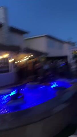 The video shows a nighttime pool party in a backyard. A group of people are gathered around a pool that is illuminated with blue lights. Some people are in the pool, some are on the edge, and some are standing around the backyard. There are inflatable rings in the pool, and balloons are visible in the background. String lights are hung around the patio area. The sky is dark, indicating it is evening or night. The atmosphere appears festive and relaxed.