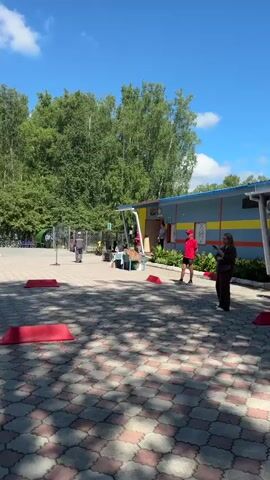 The video shows an outdoor event on a sunny day with a clear blue sky and scattered clouds. The ground is paved with interlocking hexagonal tiles in a red and grey pattern. A line of trees, primarily evergreens and birches, forms the background. In the foreground, several red, rectangular obstacles are placed on the ground.

Several people are present. Towards the center of the frame, a group of children and adults appear to be participating in or observing an activity. Some children are wearing brightly colored clothing. There are red cones scattered around the area, some of which appear to be part of an obstacle course or game.

In the background, there are various structures, including a colorful playground-like structure with slides and ramps, and a small, light-colored tent or canopy. A trailer with a wheel is also visible. Some people are standing and watching, while others are actively engaged in what seems to be a children's event or sports activity. There is a general sense of a casual outdoor gathering.