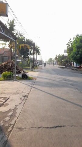 The video shows a point-of-view recording from a scooter in Kalisari, Indonesia, likely during the morning given the long shadows and bright sky. The road is paved and appears to be a moderately busy street with occasional vehicles, including motorcycles, visible in the distance. The left side of the road features trees, utility poles, and residential buildings, suggesting a suburban or semi-urban setting.

The camera, mounted on the scooter, focuses on the road ahead and the immediate surroundings. Some debris, possibly construction materials like wooden planks, is piled on the roadside. There are also hints of commercial activity with what appear to be shop signs or billboards visible in the background.

The scooter's dashboard is partially visible, showing a speedometer with red and white markings, and a Honda logo. A plastic bag containing items, possibly snacks or other small purchases, is attached to the handlebars, adding a personal touch to the scene.

The overall mood is one of everyday commuting or travel, with no indication of any specific event. The lighting suggests clear weather, and the long shadows point to either early morning or late afternoon. The scene is typical of daily life in a developing urban environment in Indonesia.